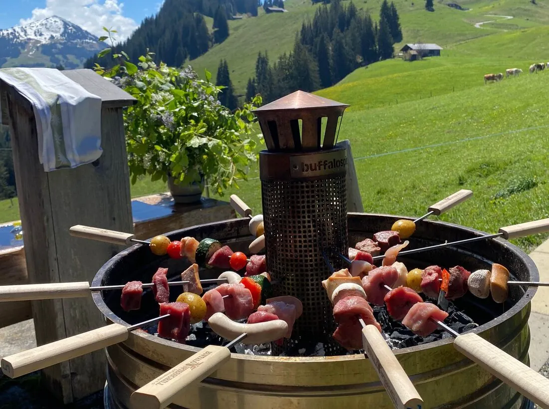 Buffalo-Spiessgrill mit aufgesteckten Fleischspiessen über der Glut, dahinter die Simmentaler Bergwiesen